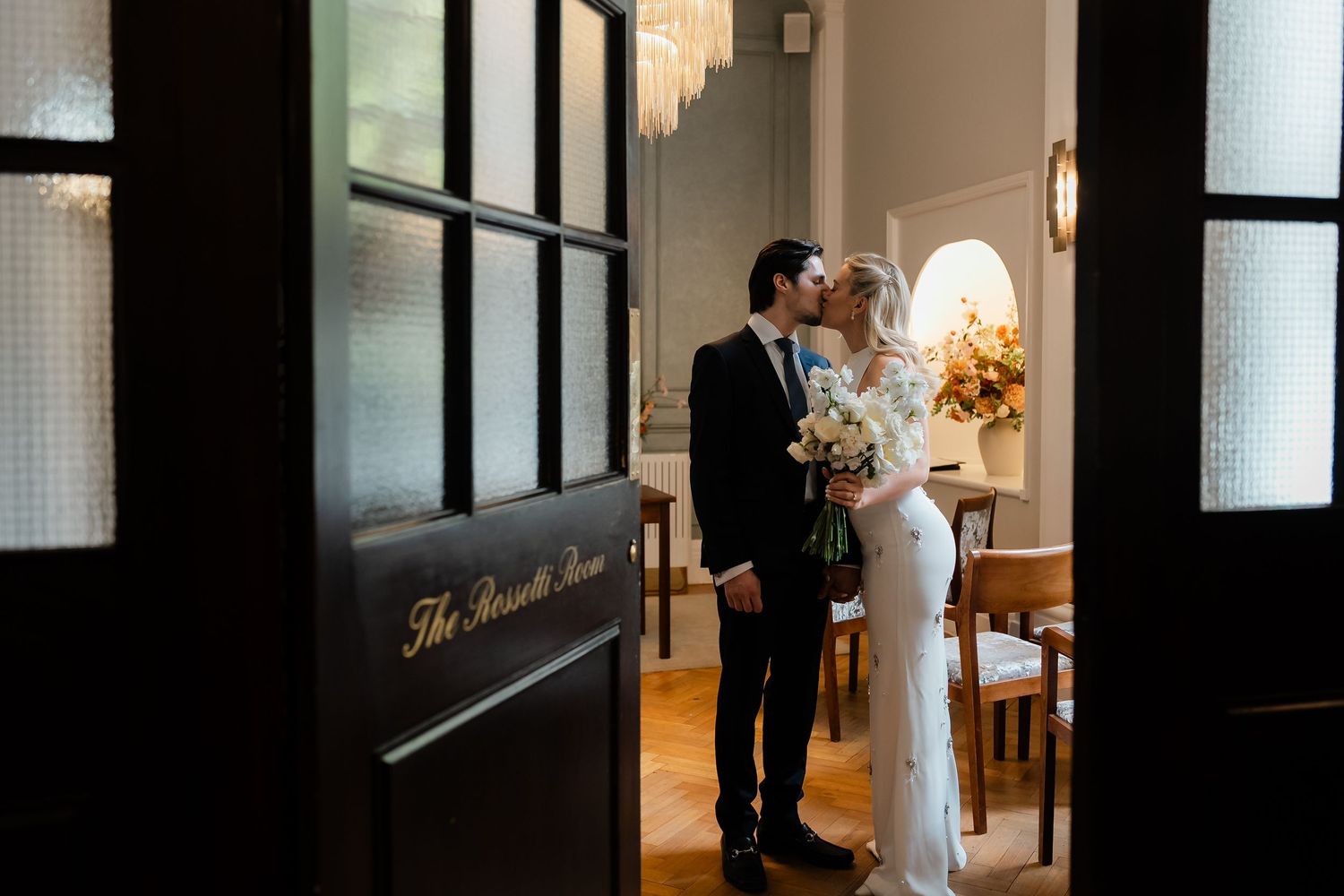 Newlyweds share a tender kiss inside The Rossetti Room, framed by elegant dark wooden doors.