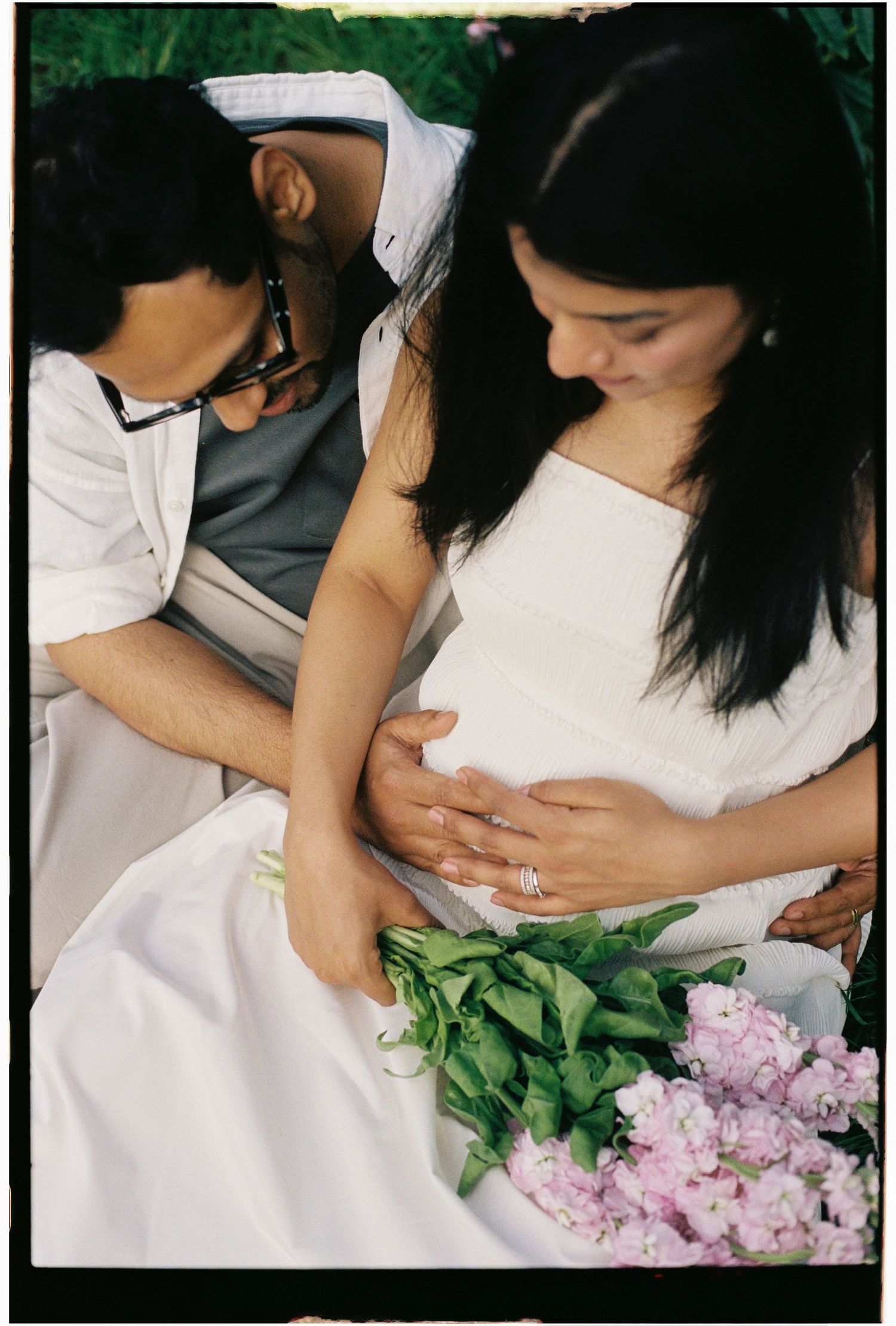 Expecting couple tenderly cradling baby bump surrounded by fresh flowers in a romantic outdoor setting.