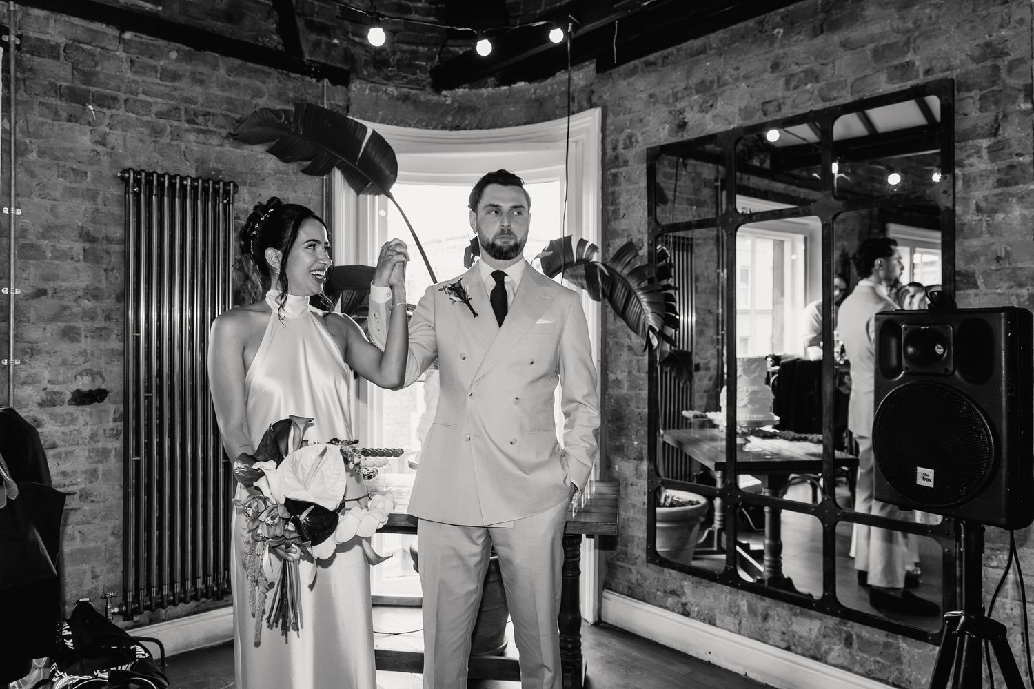 Bride and groom pose with a vintage microphone in a stylish brick-walled wedding venue.