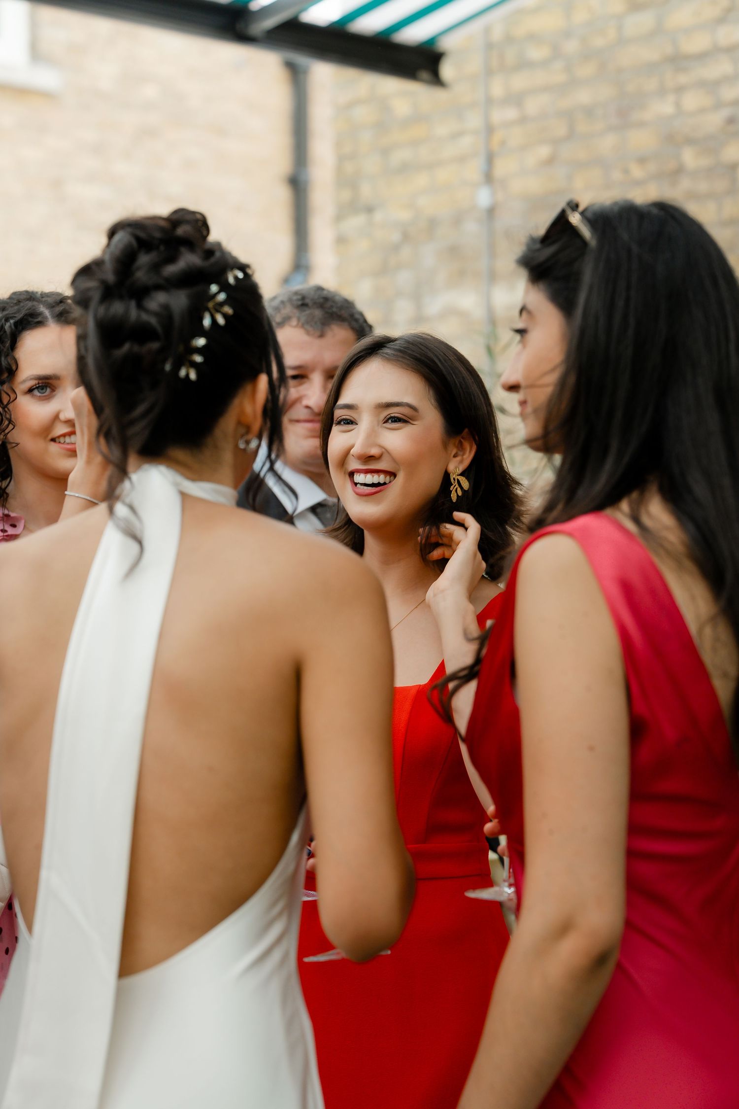Bride in a halter gown chatting and laughing with guests in red dresses at an outdoor reception.