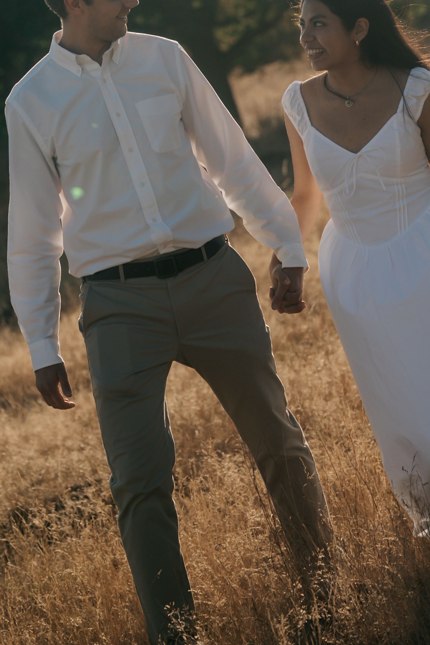 Couple walking hand in hand through golden dry grass field in warm sunset light, woman in white dress.
