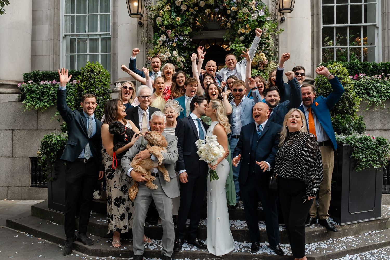 Joyful wedding group photo with guests cheering outside an elegant building entrance.