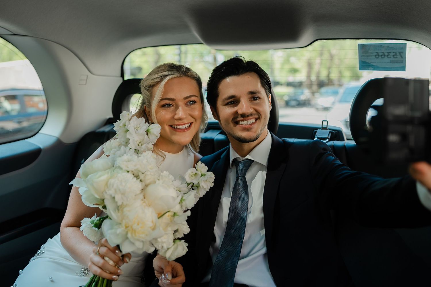 Smiling bride and groom pose together in a car backseat, bride holding a white floral bouquet.