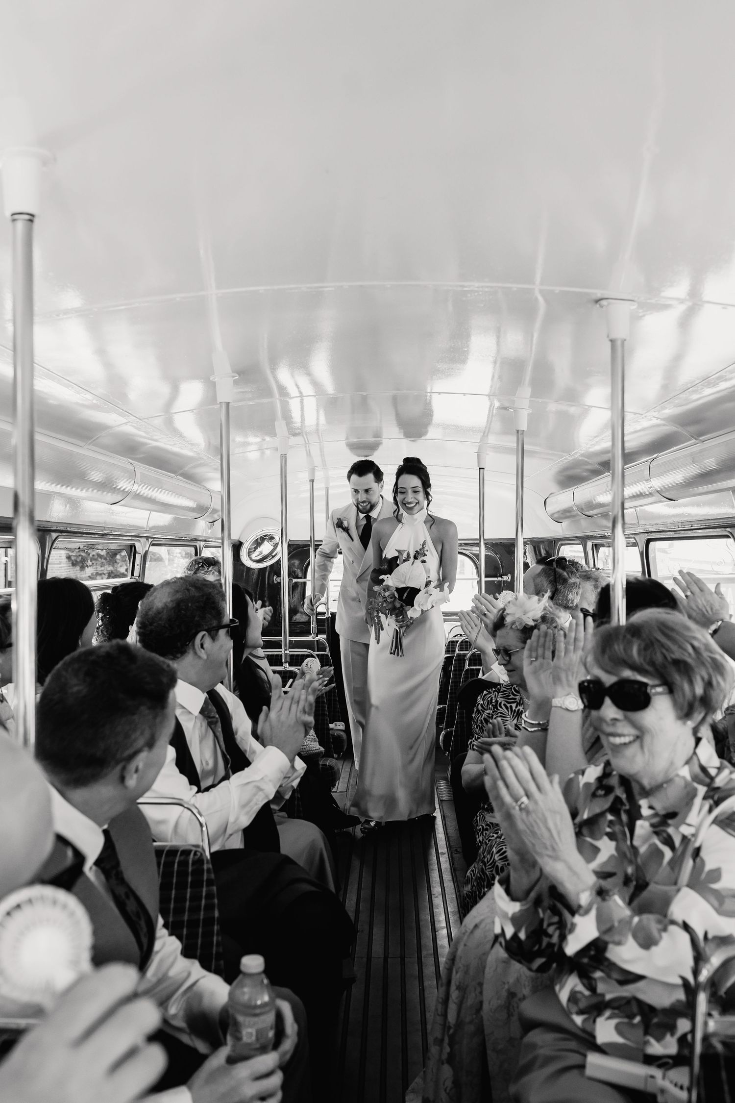 Black and white photo of bride and groom walking down the aisle of a double-decker bus as guests applaud.