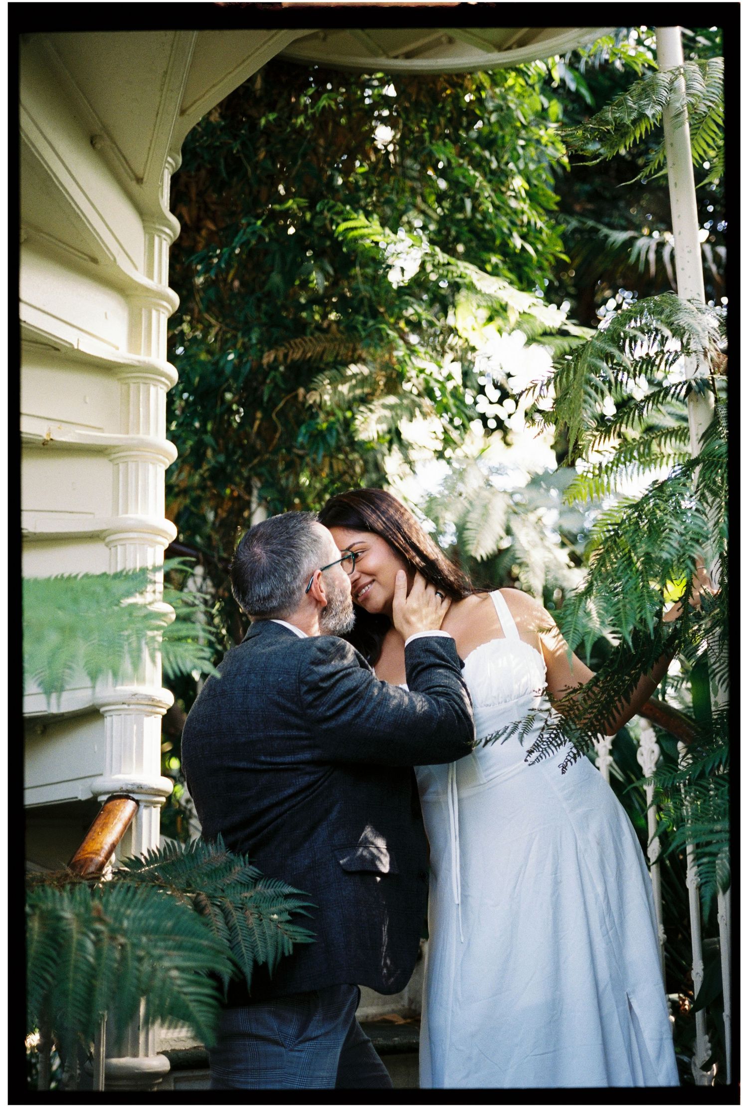 Couple sharing a romantic moment on a lush garden porch, bride in white dress and groom in dark suit.