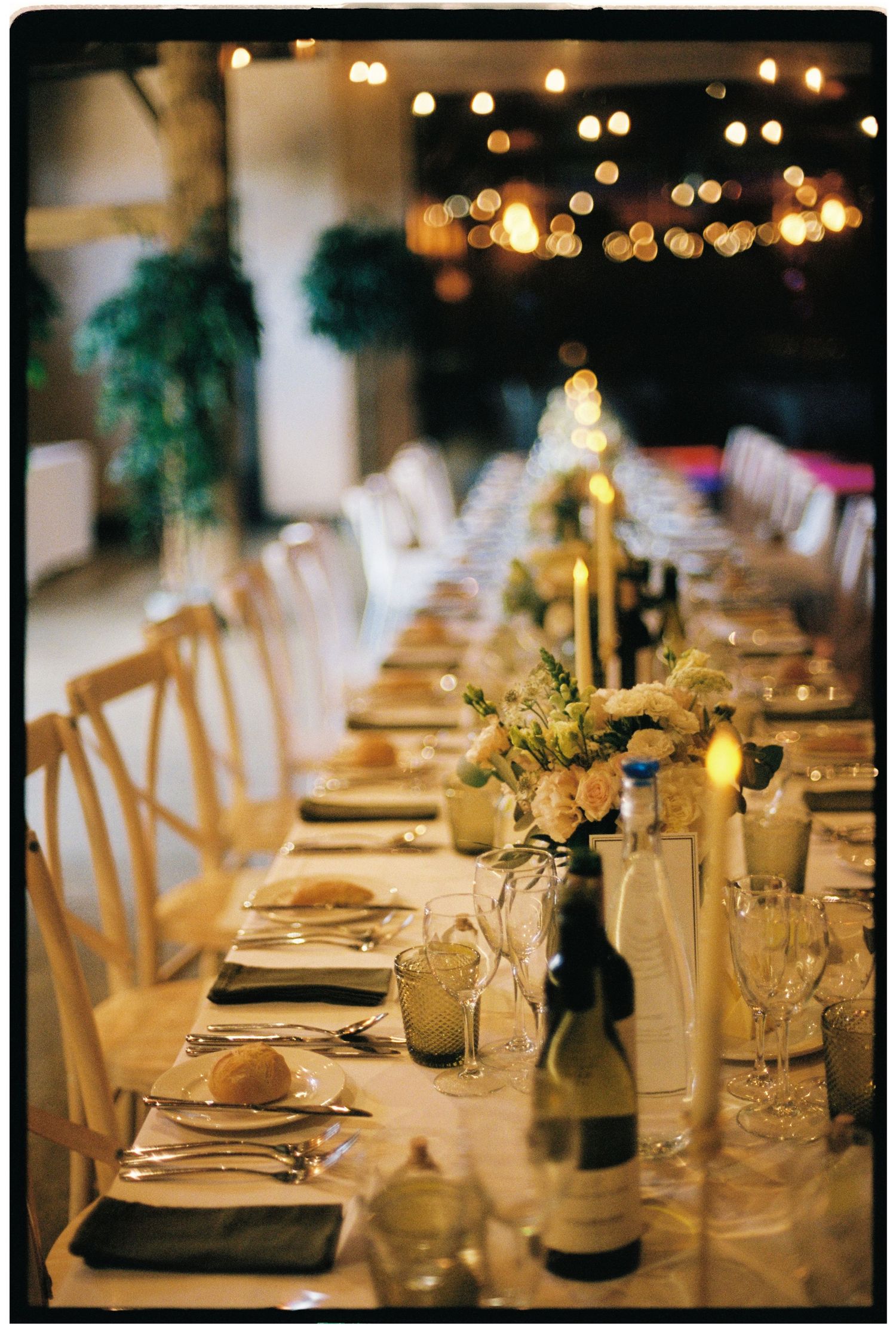 Romantic wedding reception long table setting with candles, floral centerpieces, wine bottles, and string lights.