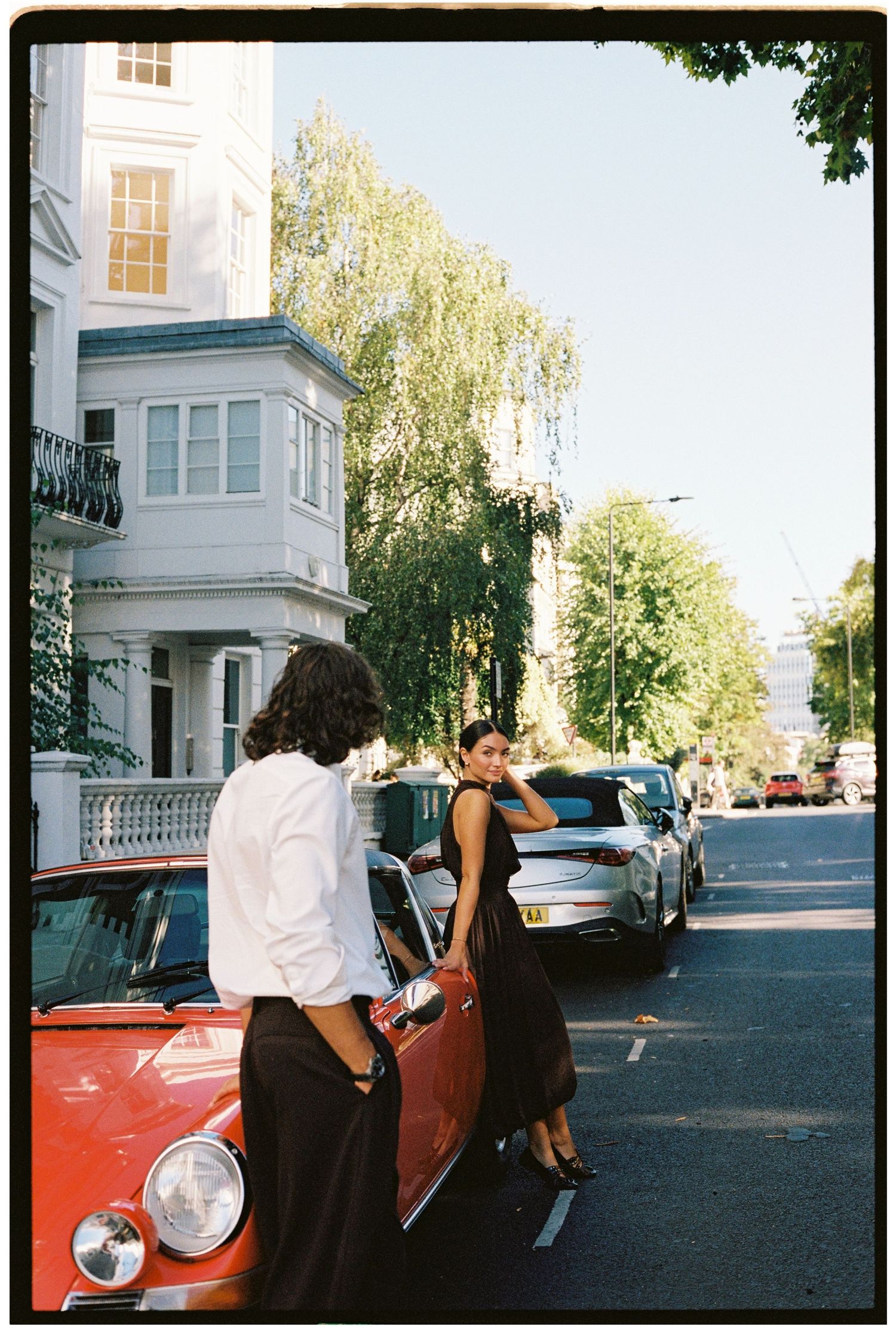 A stylish couple leaning against a red vintage car on a tree-lined street beside a white Georgian building.