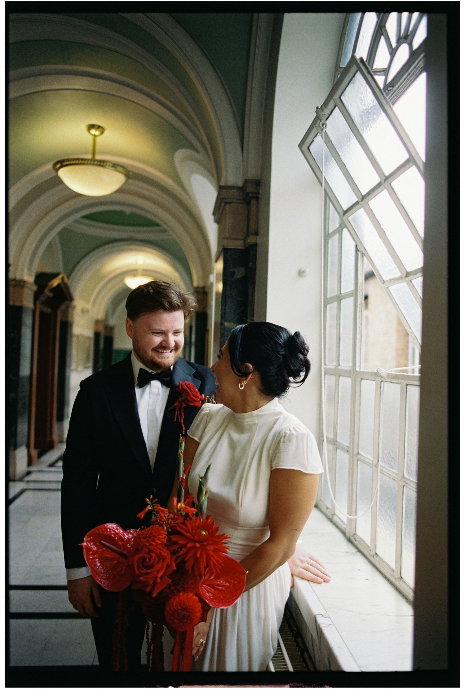 Bride and groom share a romantic moment in an elegant arched hallway, she holding a vibrant red bouquet.
