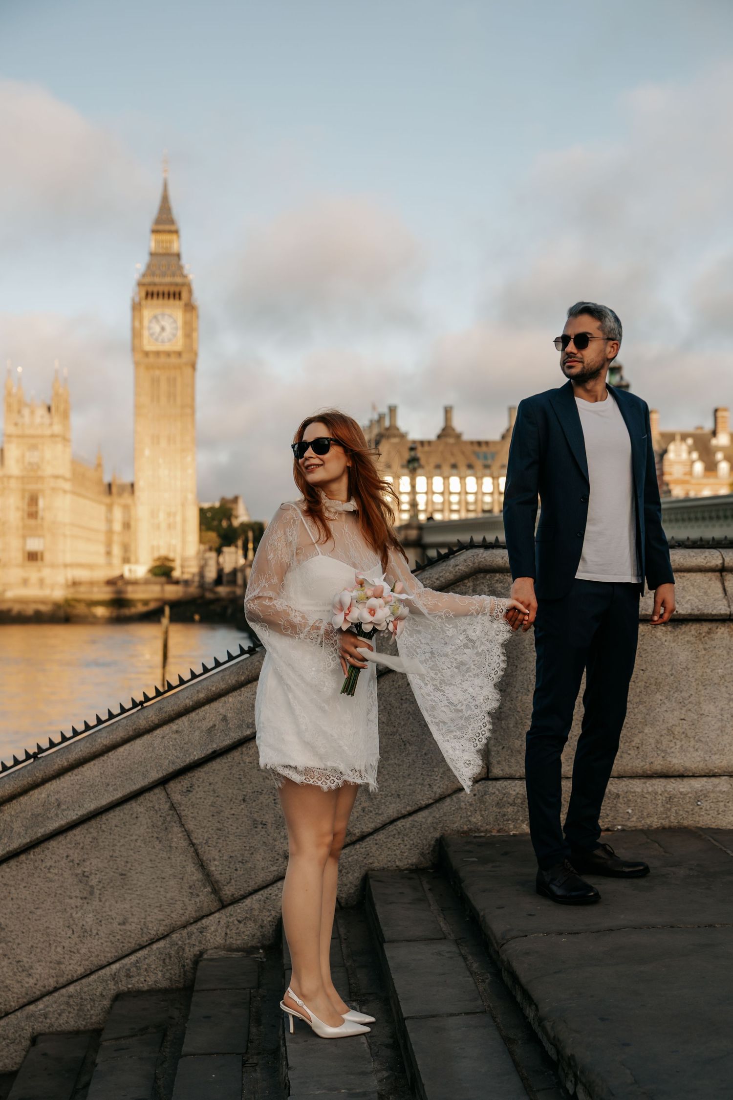 Bride and groom pose on steps near Big Ben, London, bride holding bouquet in white lace dress.