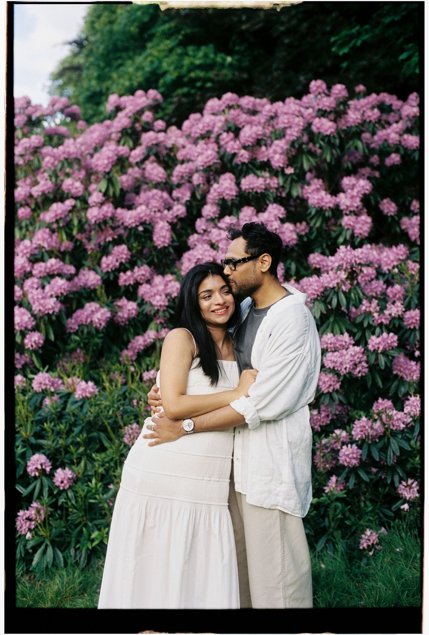 Couple embracing before pink flowering shrubs, man kissing woman's cheek, both in white outfits.