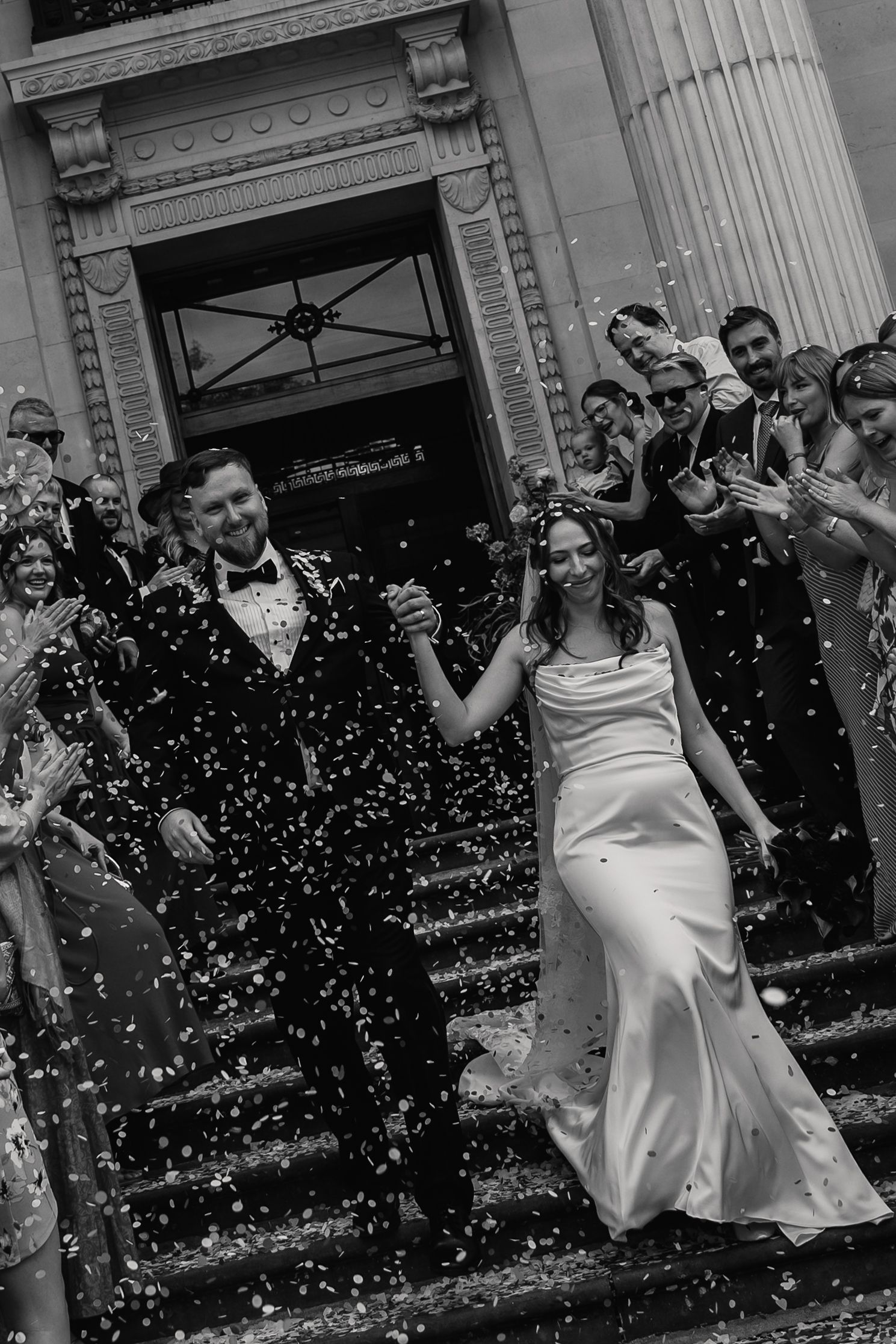 Black and white photo of smiling newlyweds descending steps through confetti with celebrating guests.