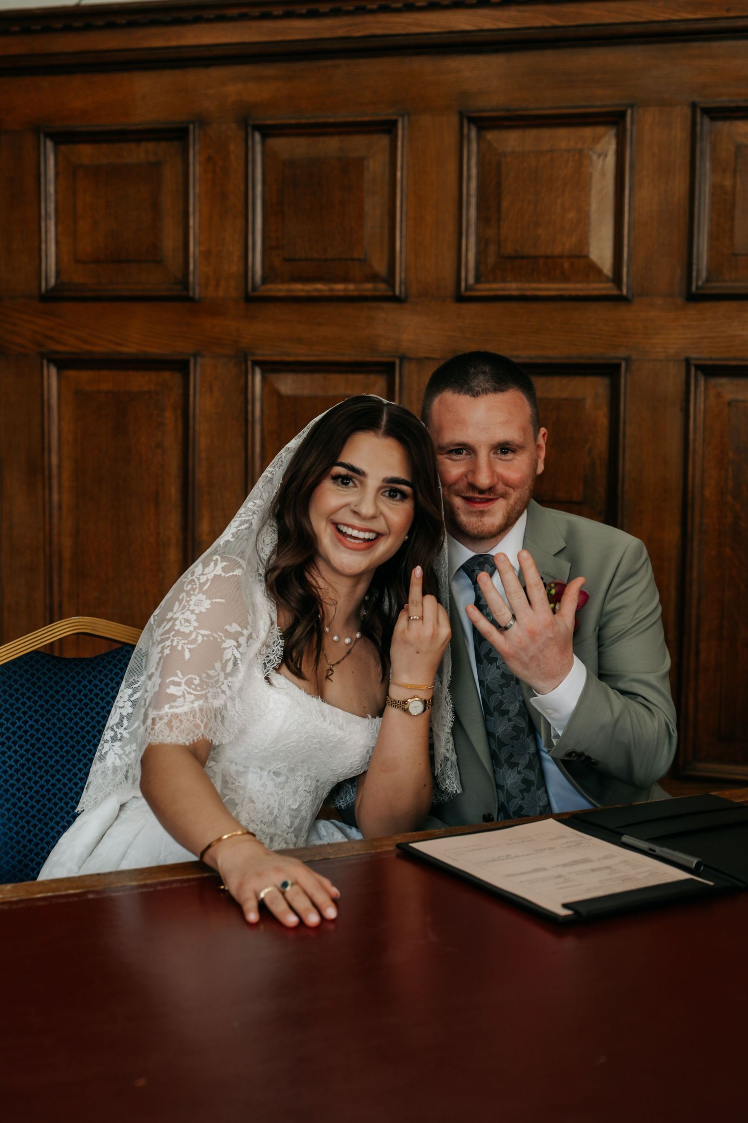 Newlyweds sitting at a table showing off their wedding rings after signing the marriage register.