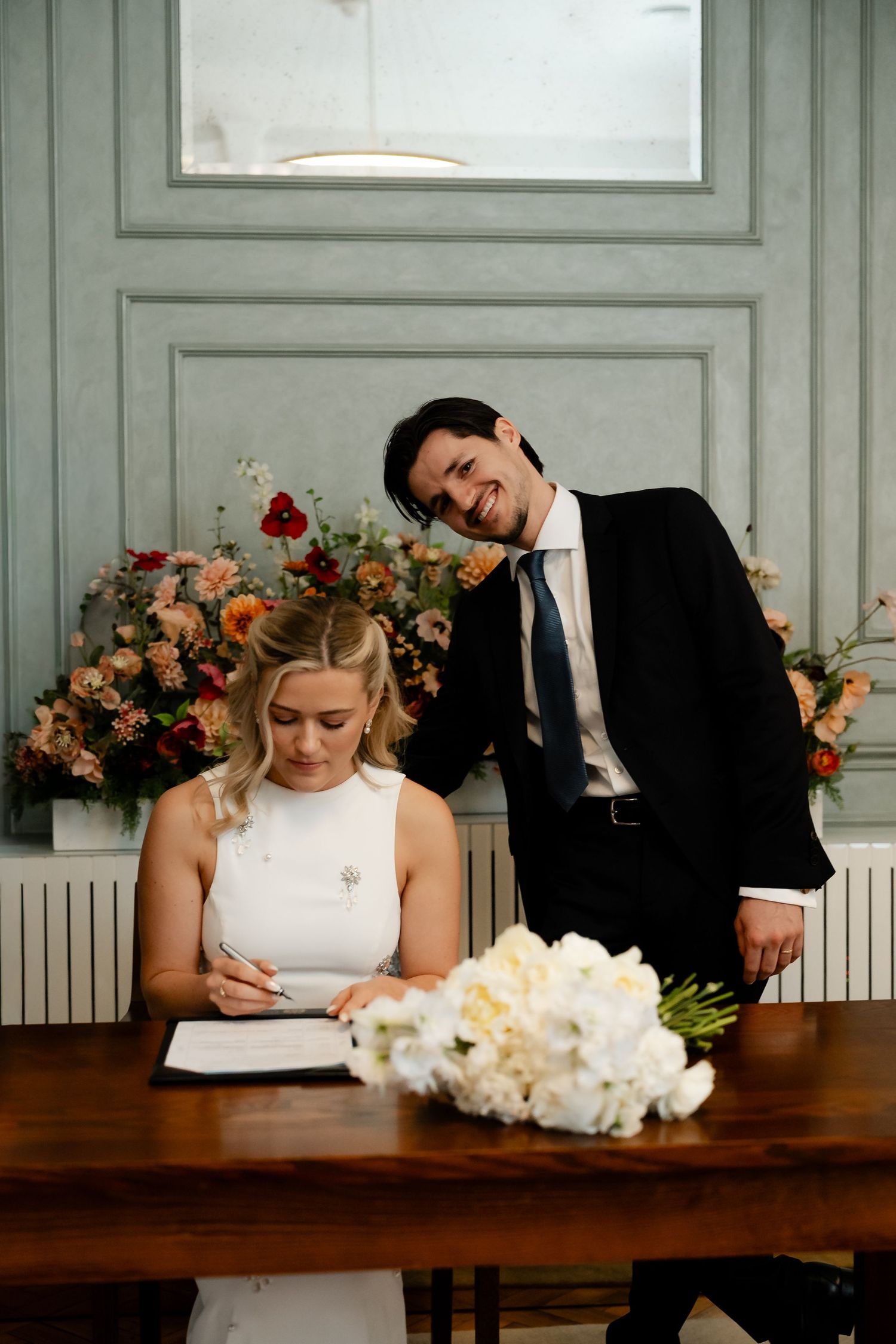 Bride signs wedding register at wooden table while groom smiles proudly beside floral arrangements.