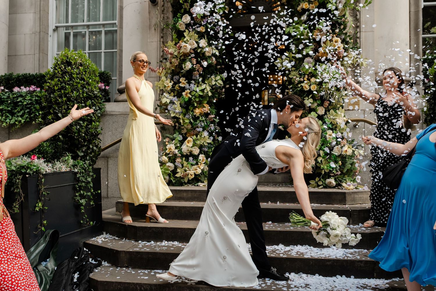 Wedding couple dips during confetti shower outside a stunning floral-decorated venue entrance.
