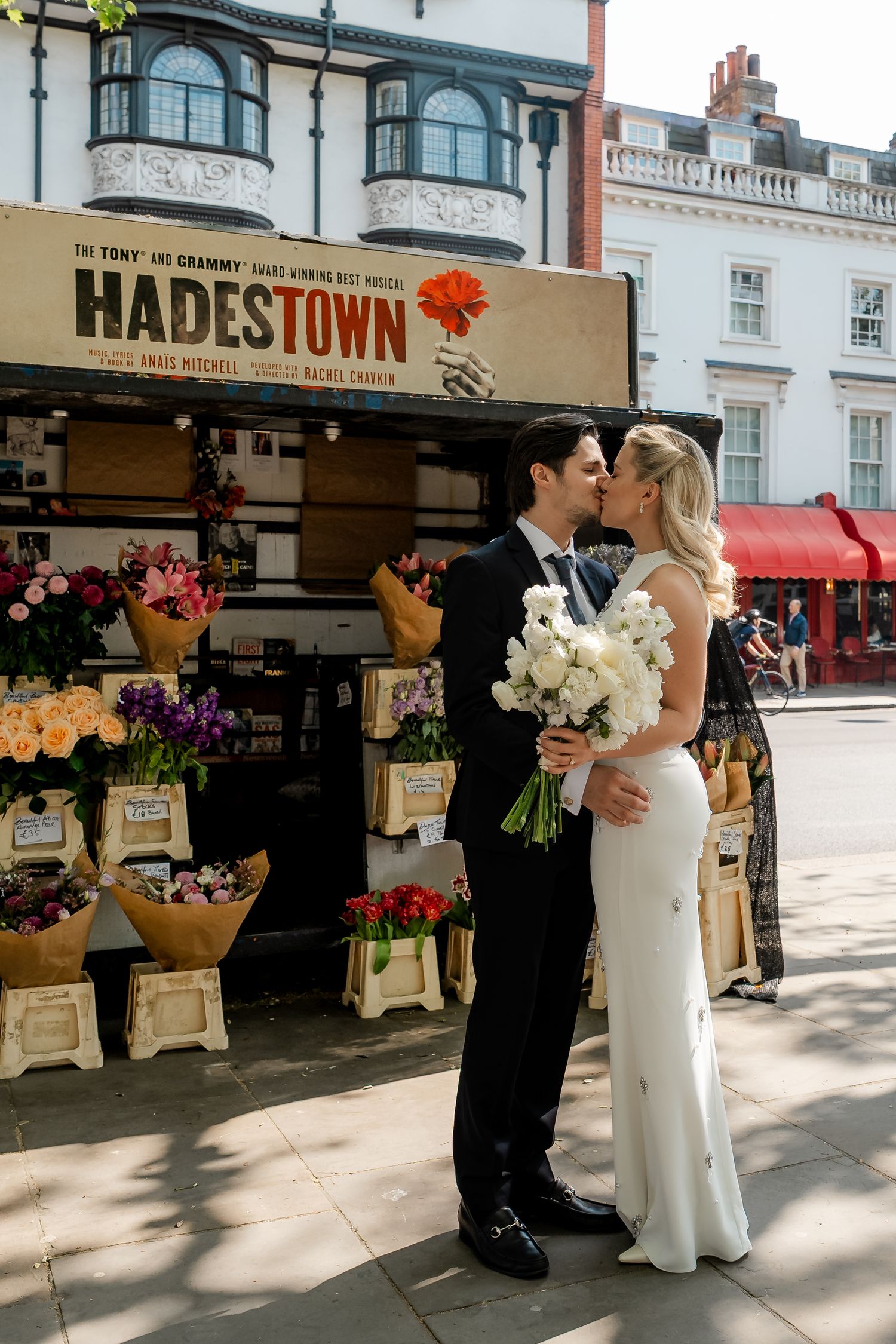 Wedding couple kissing in front of a Hadestown musical poster at a vibrant London flower market stall.