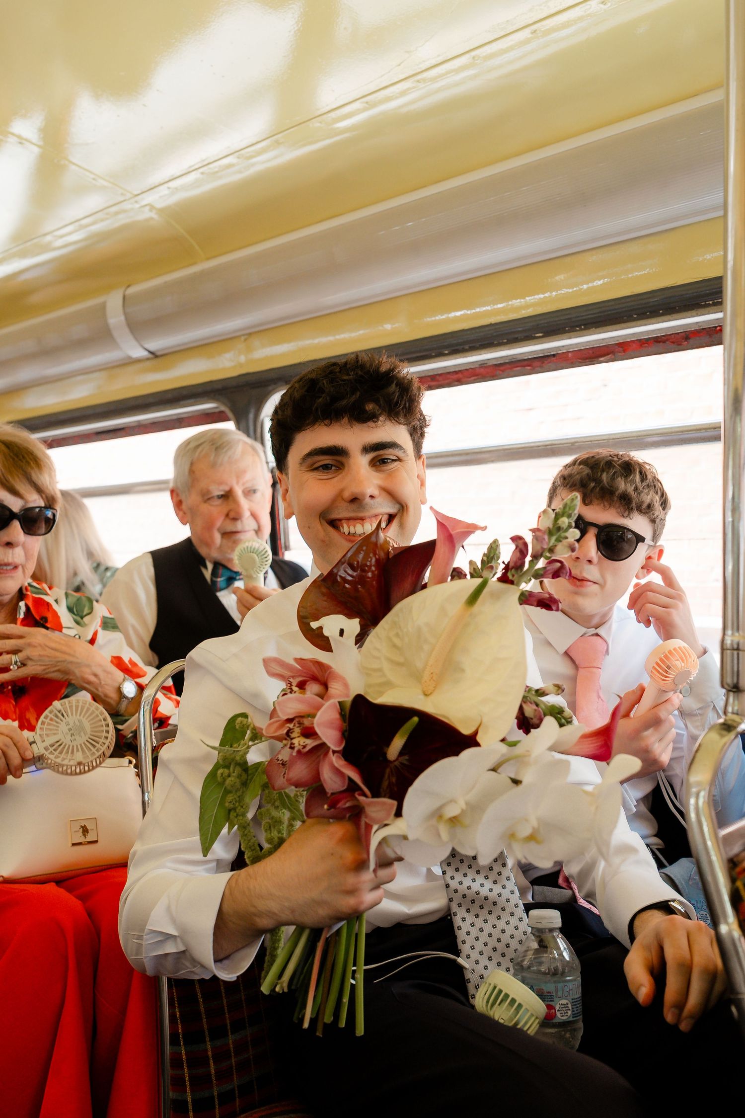 Smiling young wedding guest holding the bridal bouquet of white orchids and tropical flowers inside a vintage bus.