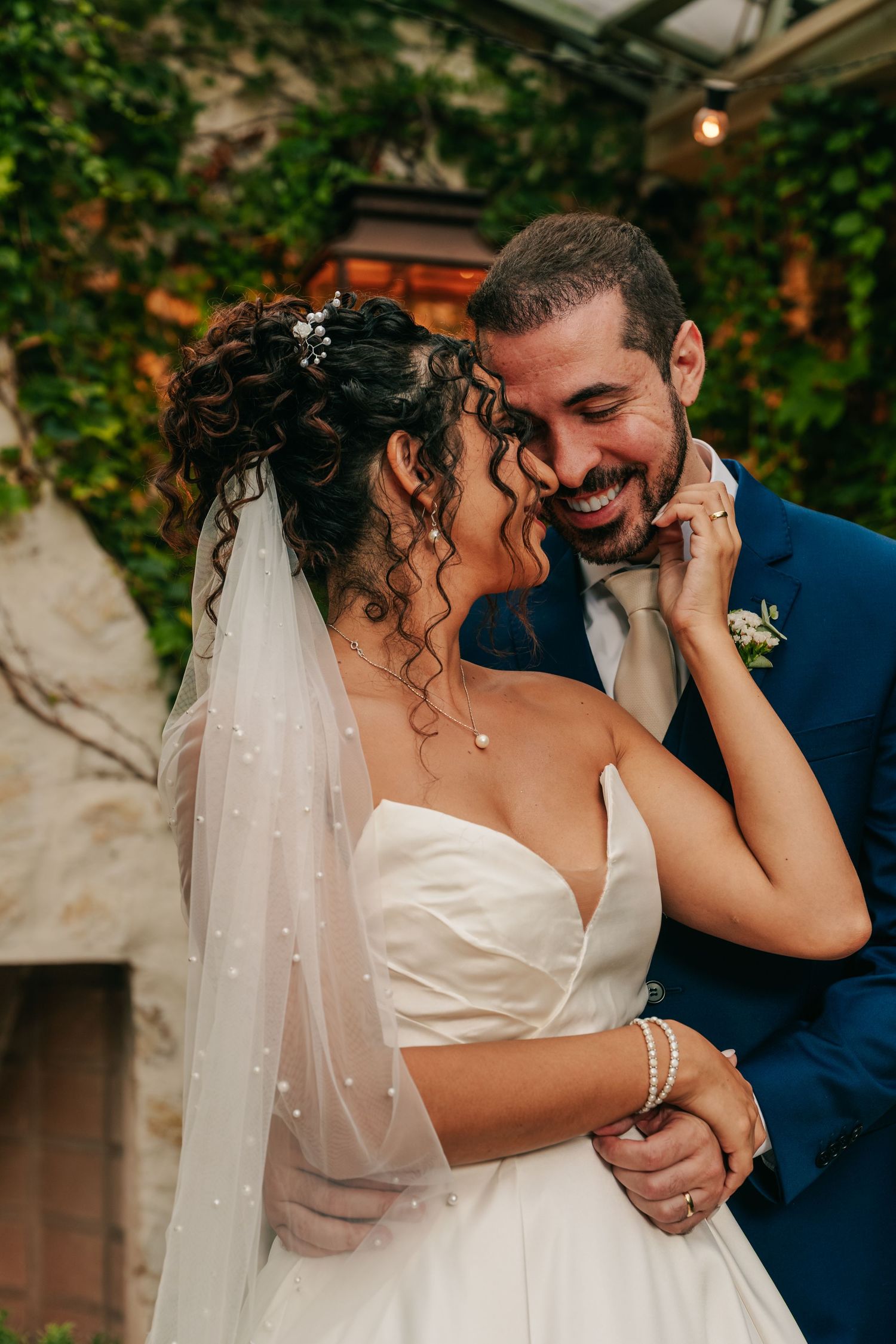 Joyful bride and groom in blue suit share intimate moment, embracing and smiling in ivy-covered outdoor venue.