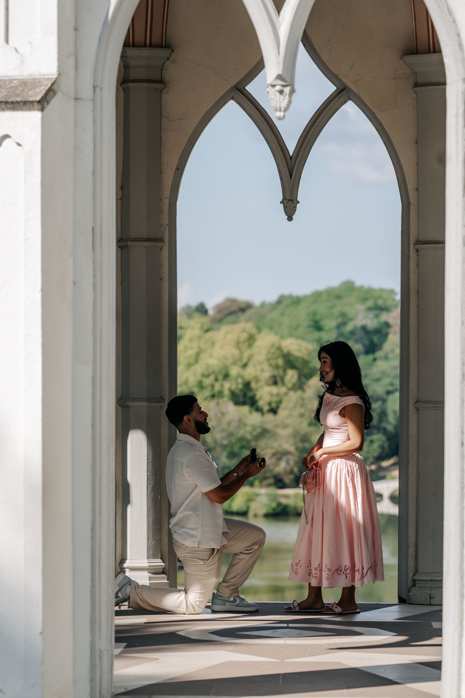 Man proposing on one knee to woman in pink dress under Gothic arch with lake backdrop.