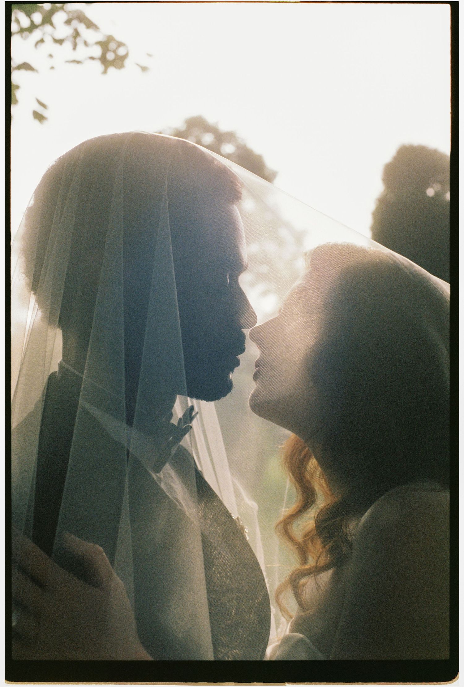Romantic backlit silhouette of bride and groom beneath a veil with golden sun flare.