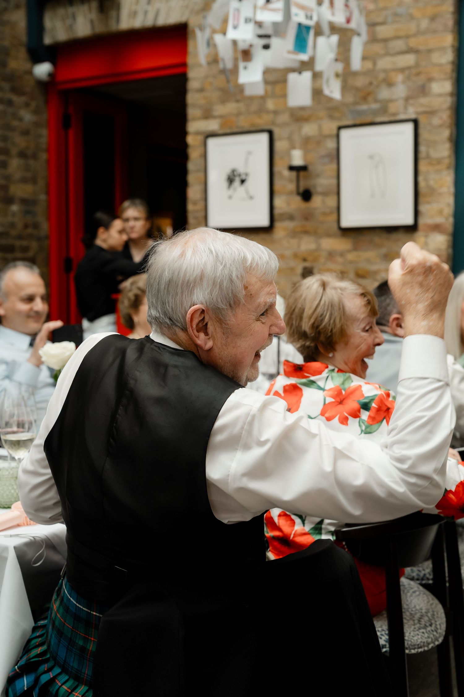Elderly man in tartan kilt laughing joyfully at wedding reception dinner table.