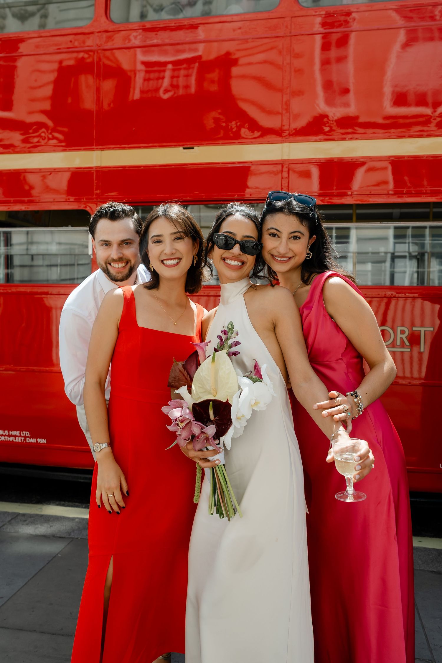 Bride in white halter gown with wedding party in red dresses posing in front of iconic red London double-decker bus.