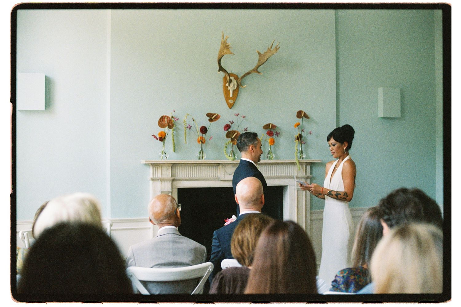 Bride reading vows at a ceremony with floral arrangements above a fireplace and seated guests.