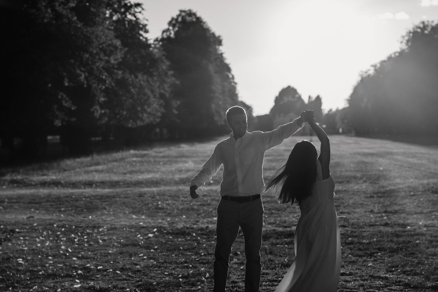 Black and white photo of couple dancing in sunlit open field surrounded by trees.