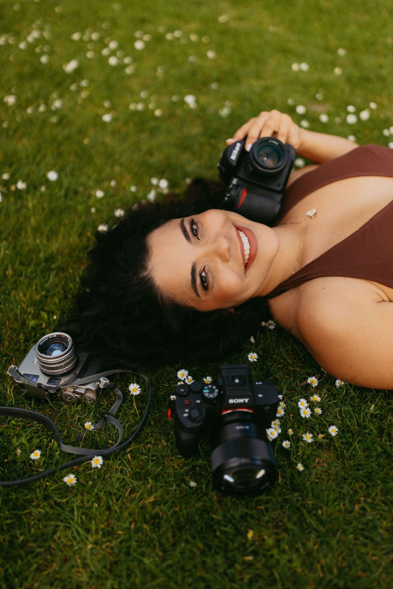 Smiling woman lying on grass holding cameras, surrounded by daisies, with Sony and vintage film cameras nearby.