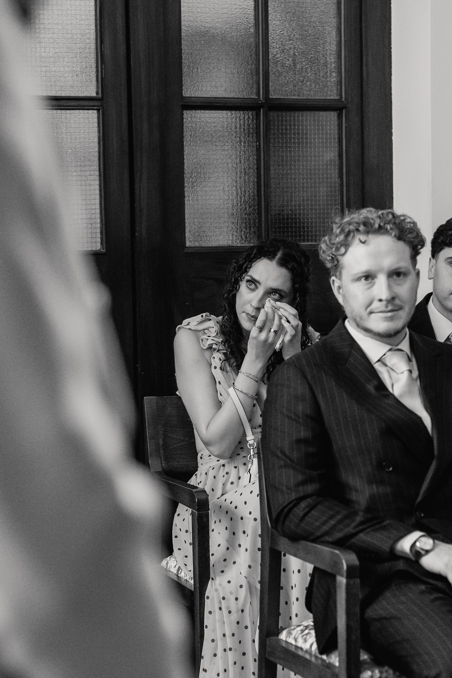 Emotional wedding guest wipes tears while watching the ceremony in a black and white candid photo.