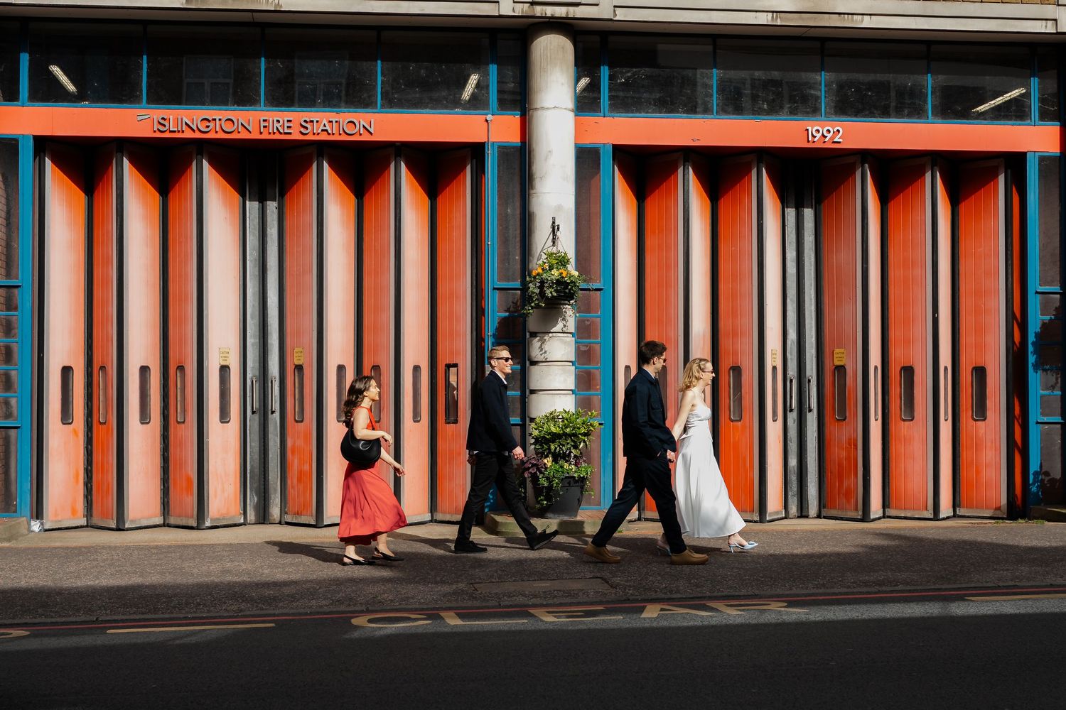 Wedding party walking past iconic red Islington fire station facade on sunny day.