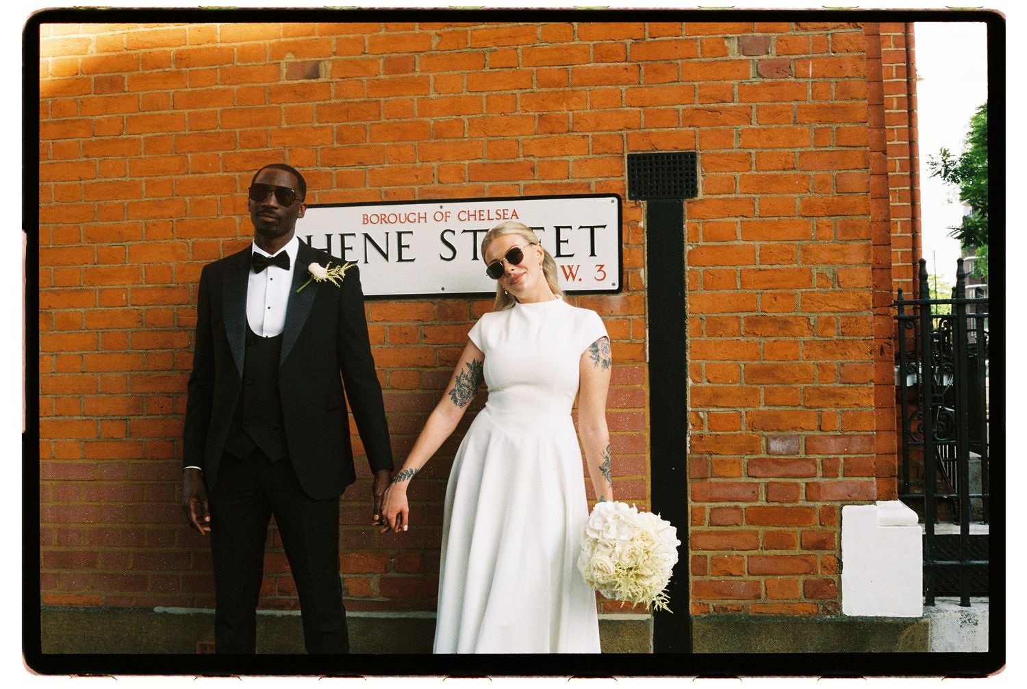 Interracial couple in wedding attire pose in front of a London street sign on brick wall.