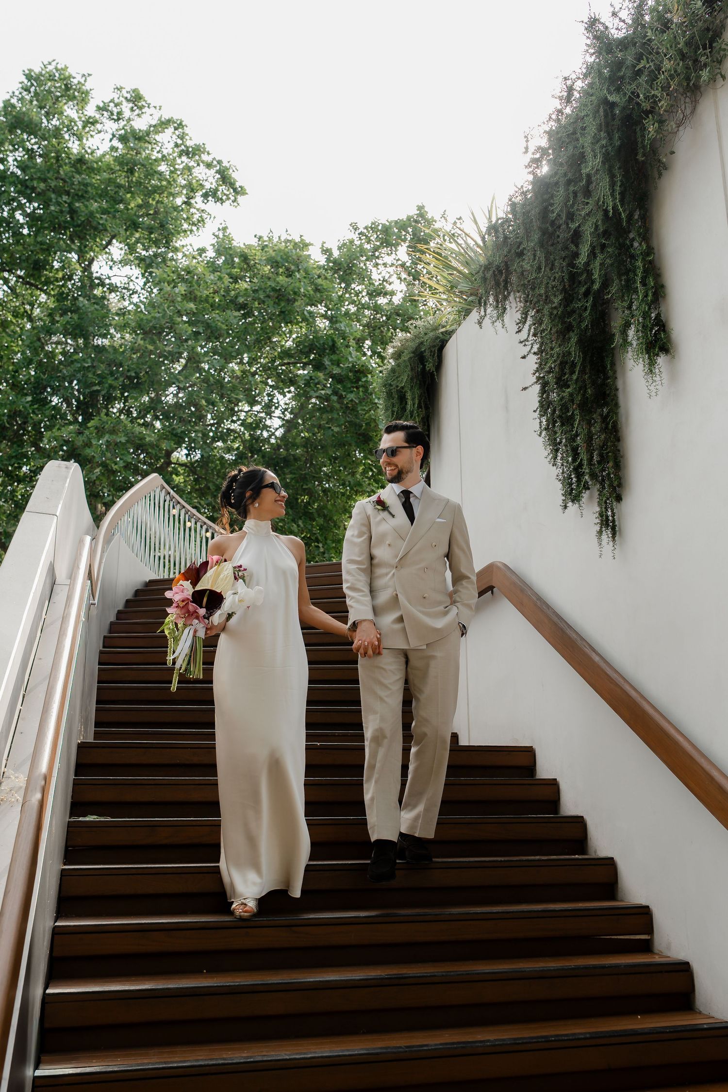 Bride and groom descending wooden staircase with lush greenery, bride holding colorful wedding bouquet.