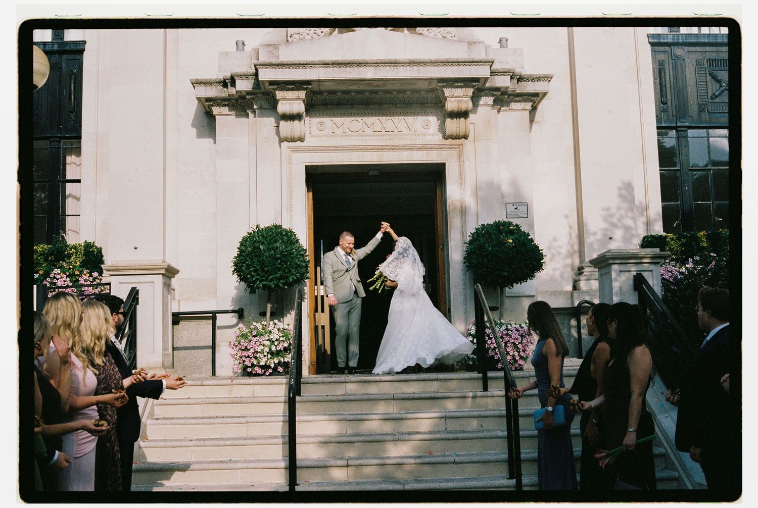 Bride and groom exit a grand church entrance, groom raising bride's hand, guests watching on steps.