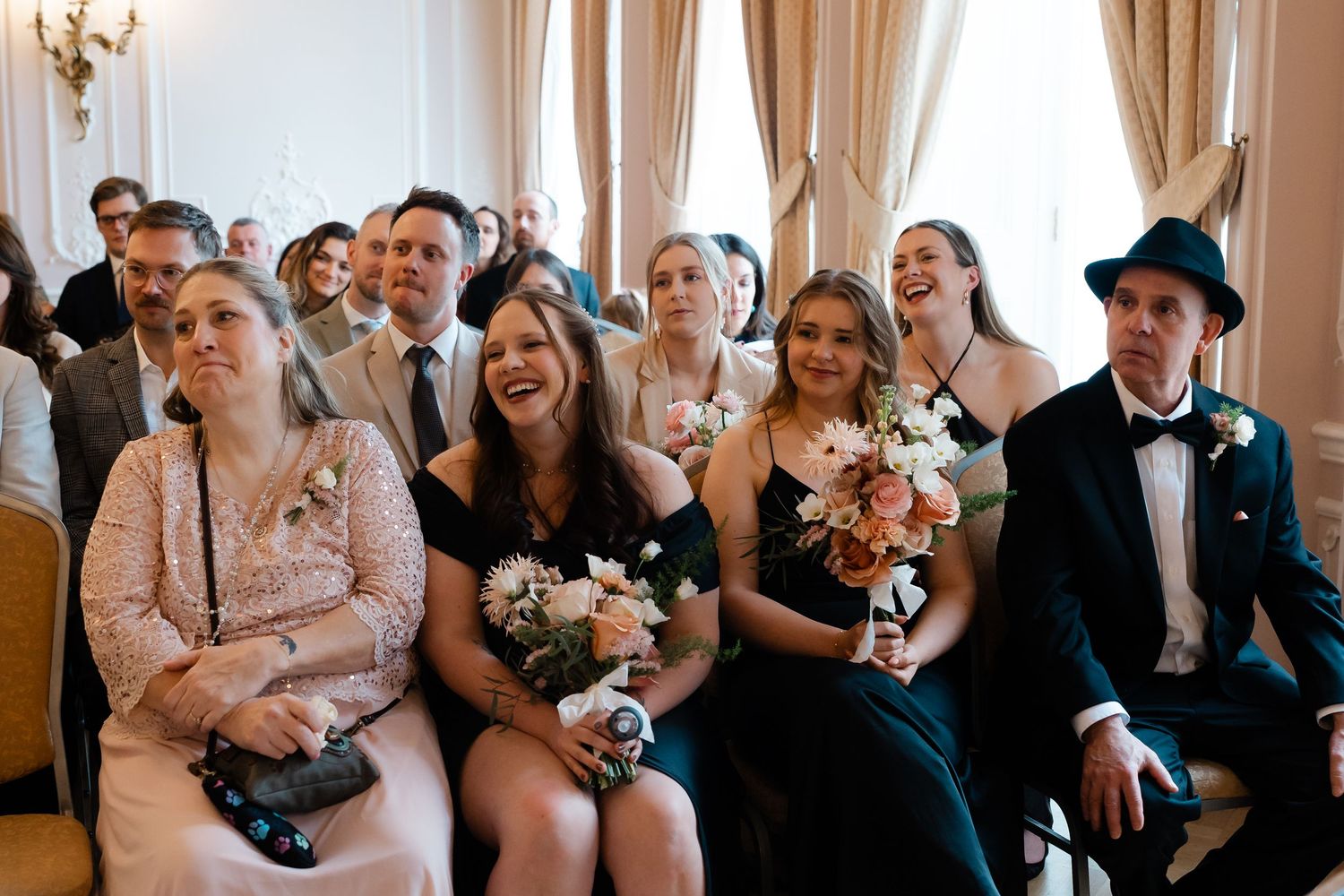 Wedding guests seated at ceremony smiling and holding bouquets in an elegant decorated room.