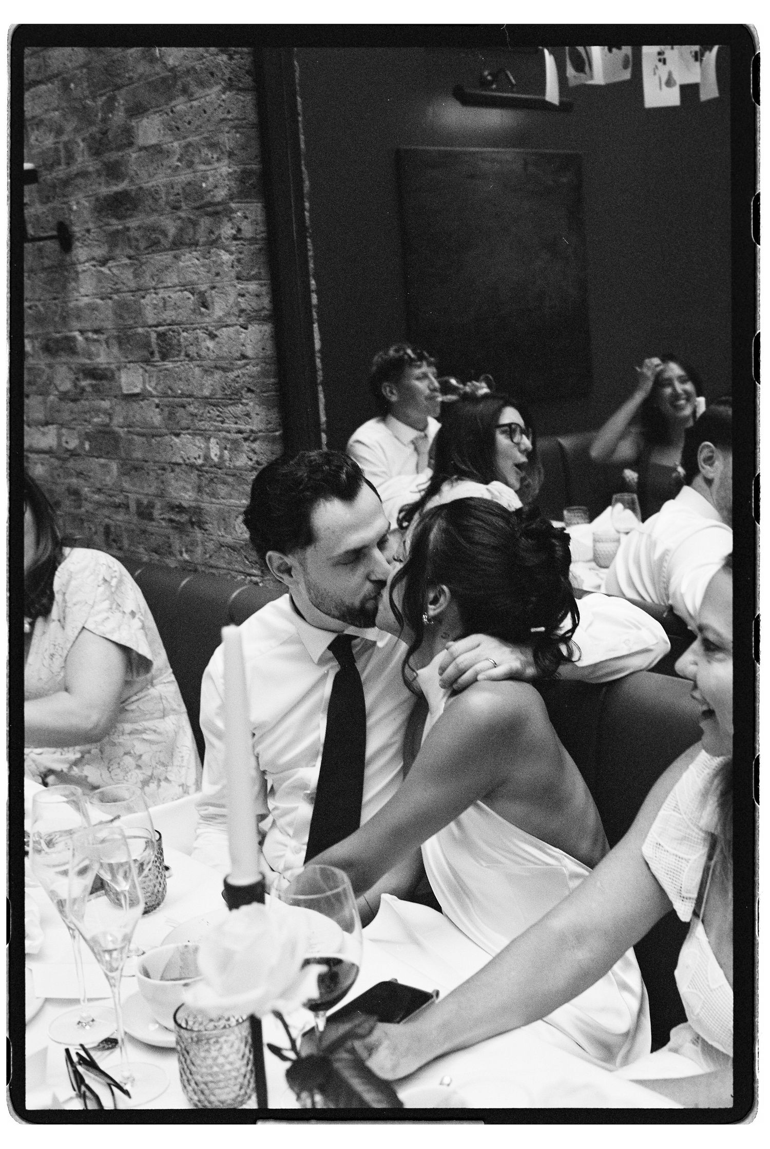 Black and white photo of bride and groom sharing a kiss at wedding reception surrounded by guests.