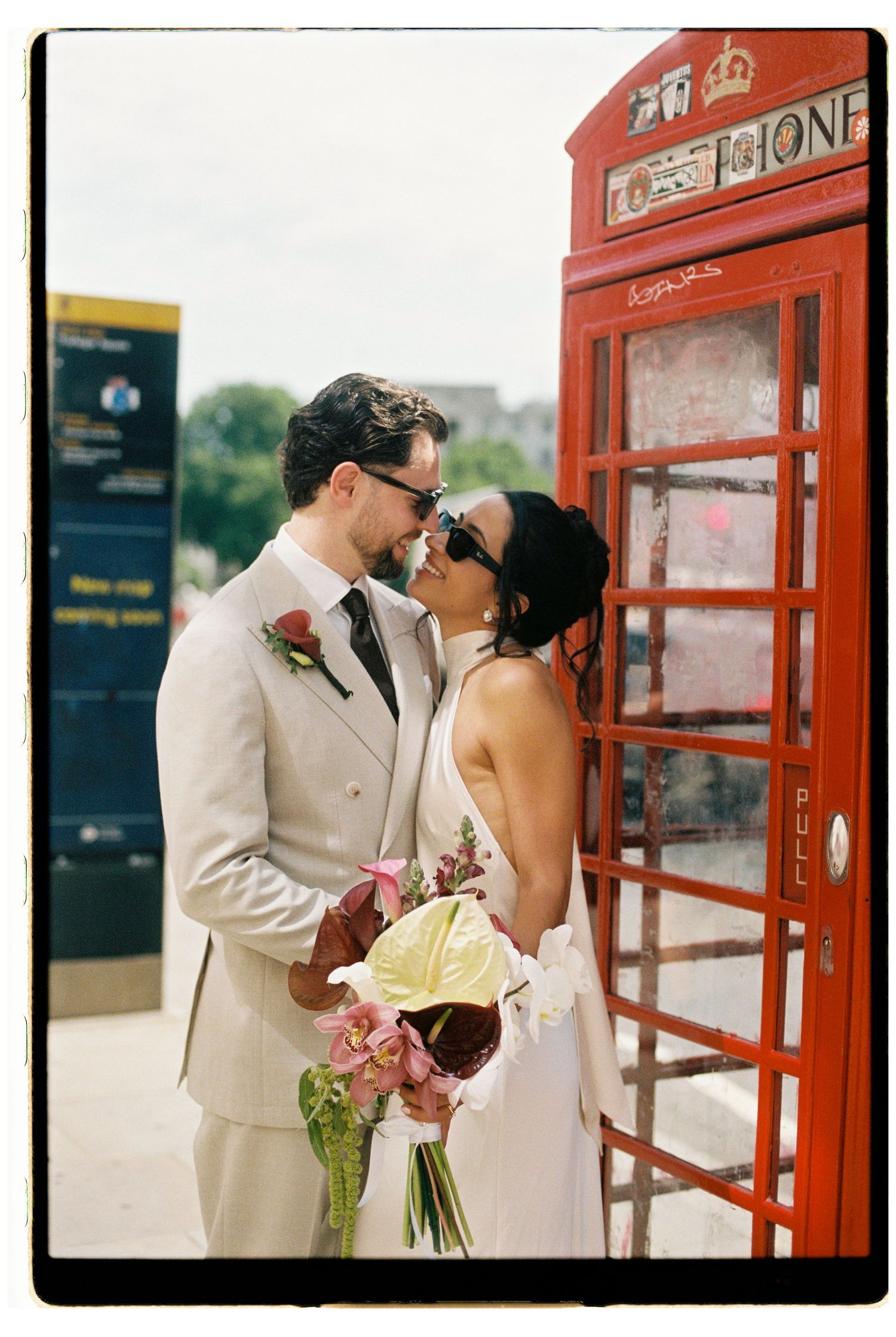 Bride and groom share a kiss beside a red London phone box in a vintage film-style photograph.