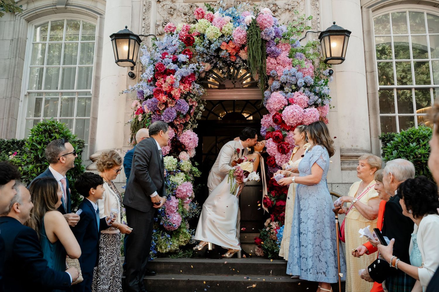 Wedding couple kissing on steps beneath vibrant pink and blue floral arch.