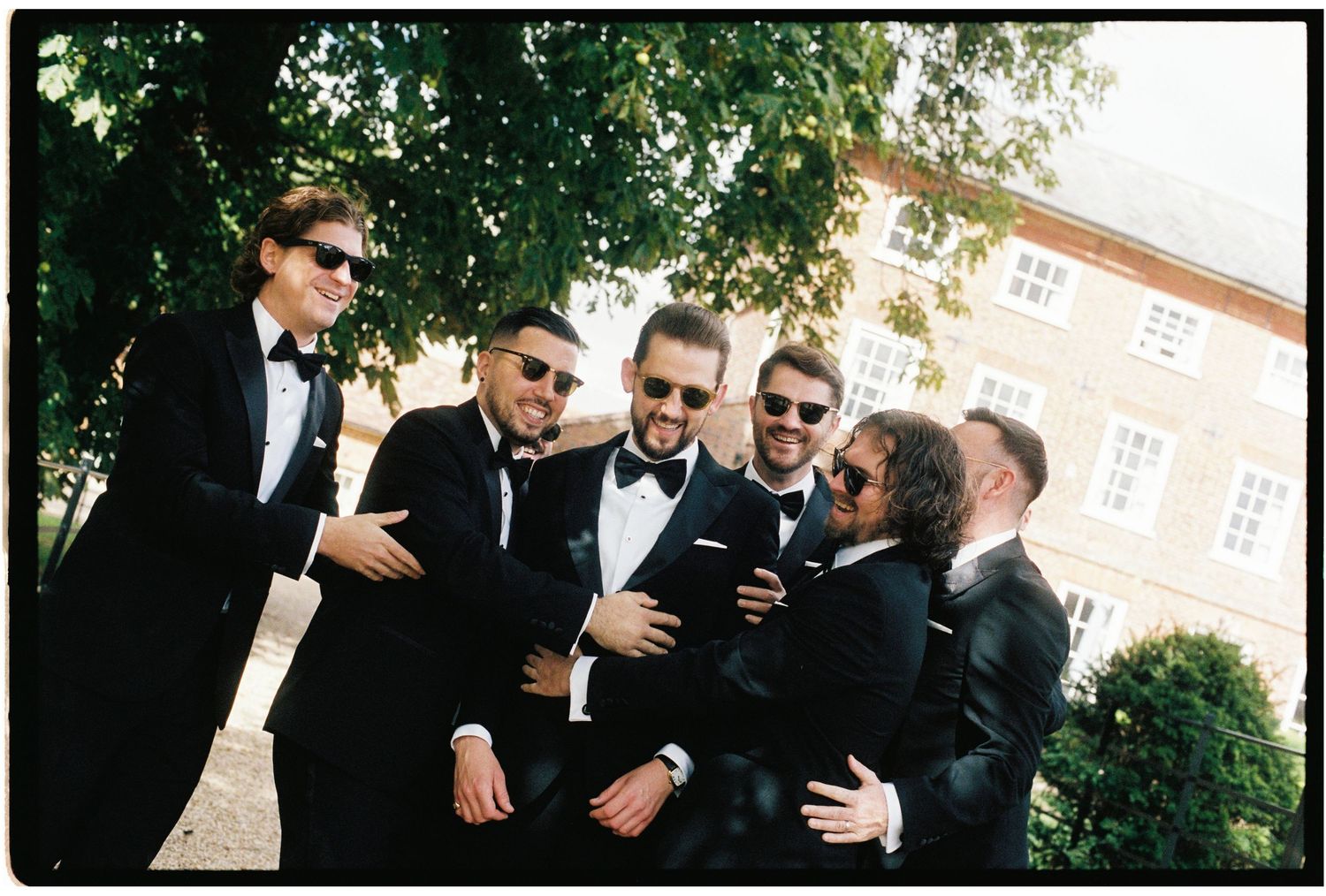 Group of groomsmen in black tuxedos laughing together outdoors near a historic building.