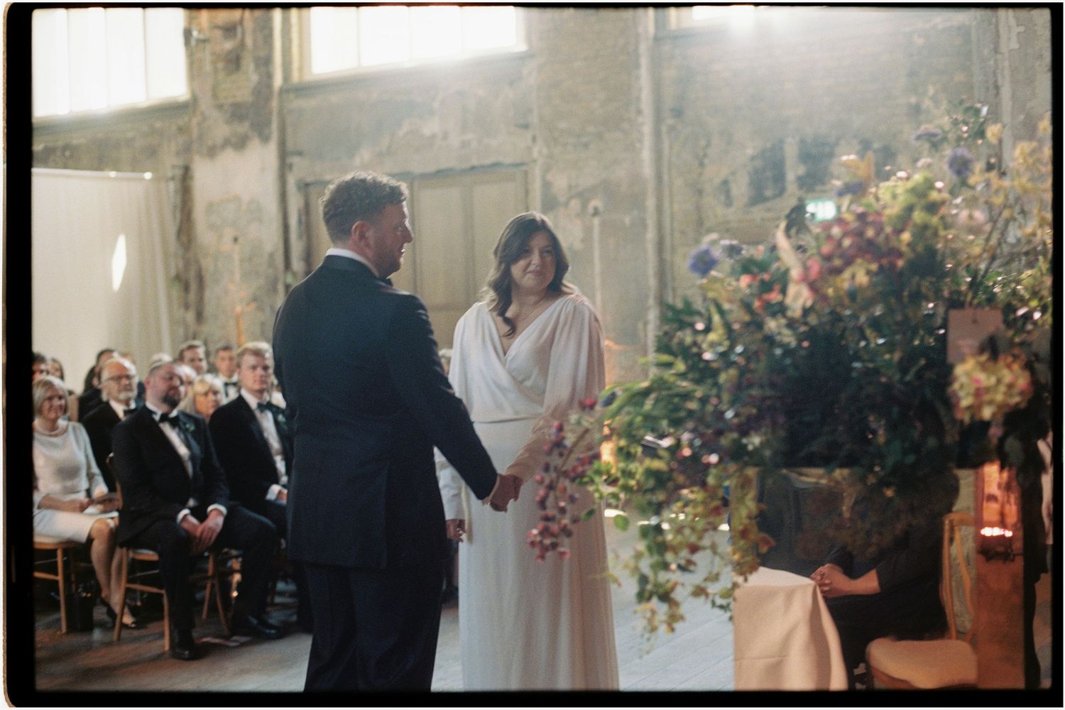 Bride and groom hold hands during intimate wedding ceremony with floral arrangement and seated guests in background.