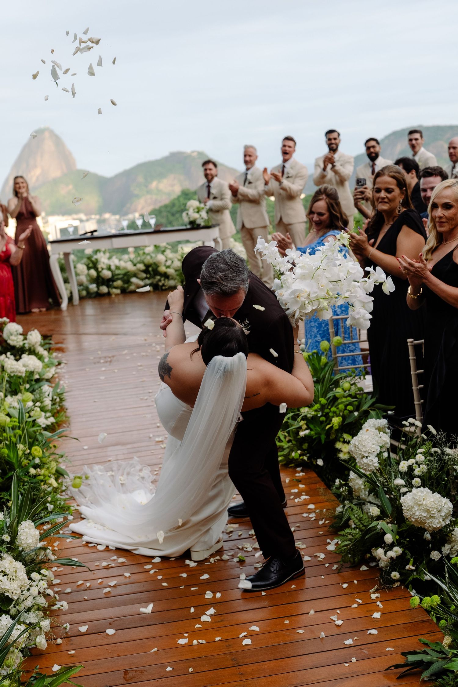 Groom dips bride for kiss as guests celebrate with flower petals at outdoor Rio wedding ceremony.