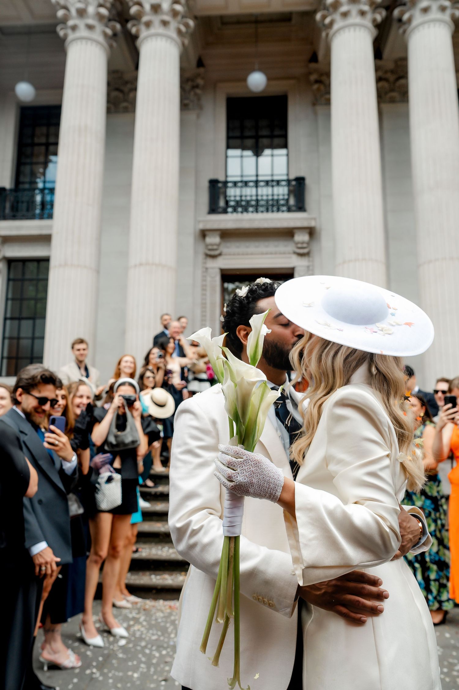 Couple sharing a kiss outside a grand columned building on their wedding day, bride wearing a white hat and groom in a white suit.