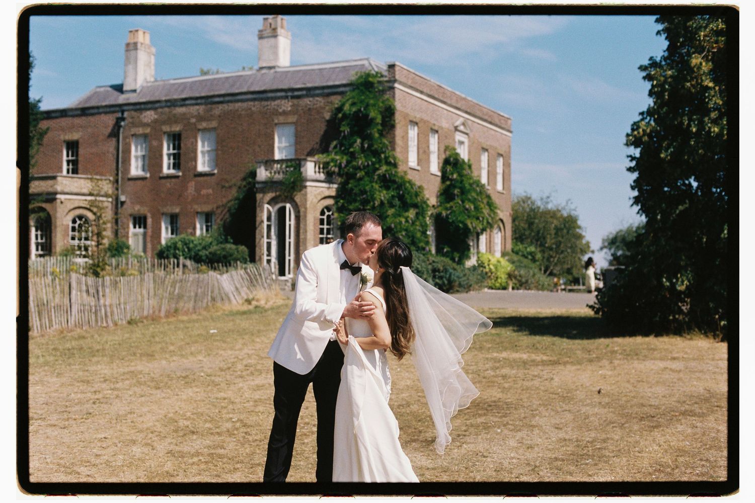 Couple sharing a kiss in front of a grand Georgian manor house on a sunny wedding day.