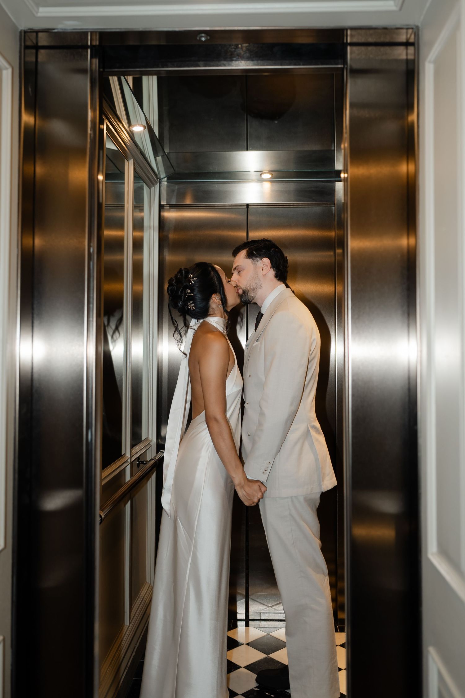 Newlyweds sharing a romantic kiss while holding hands inside a sleek metallic elevator on their wedding day.
