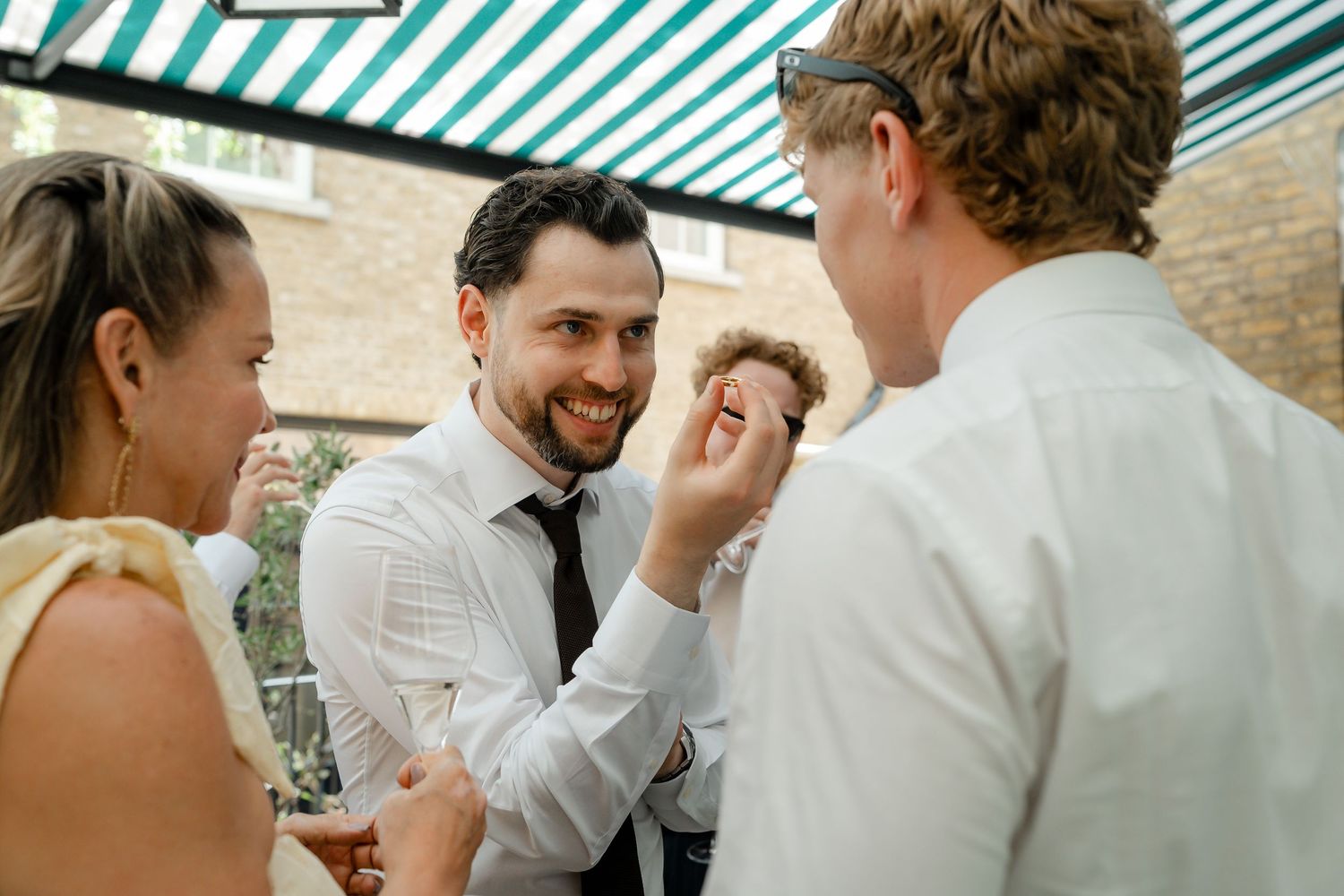 Smiling groom in a white shirt and tie laughing with guests at an outdoor wedding reception.