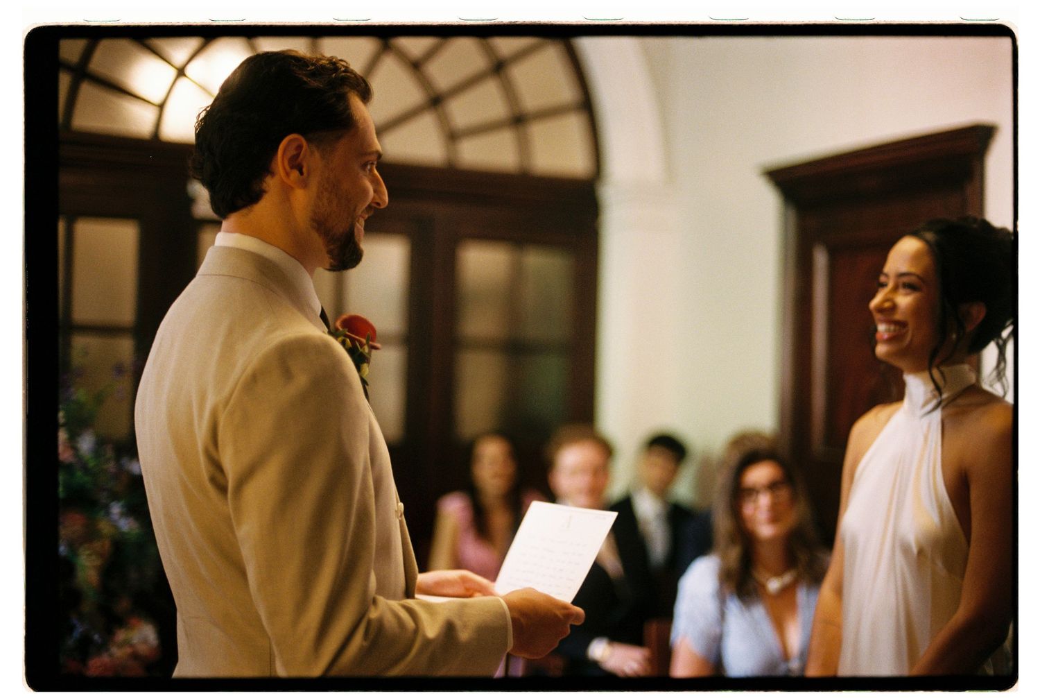 Groom in beige suit reading vows to smiling bride at intimate wedding ceremony.
