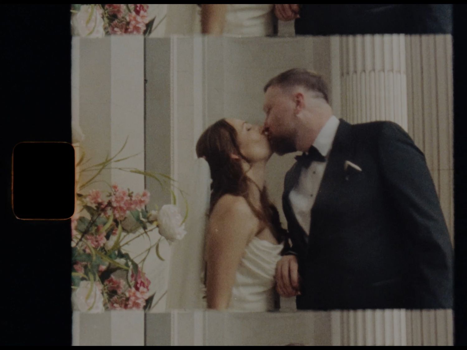 A bride and groom share a romantic kiss at their wedding ceremony, surrounded by floral arrangements and elegant columns.