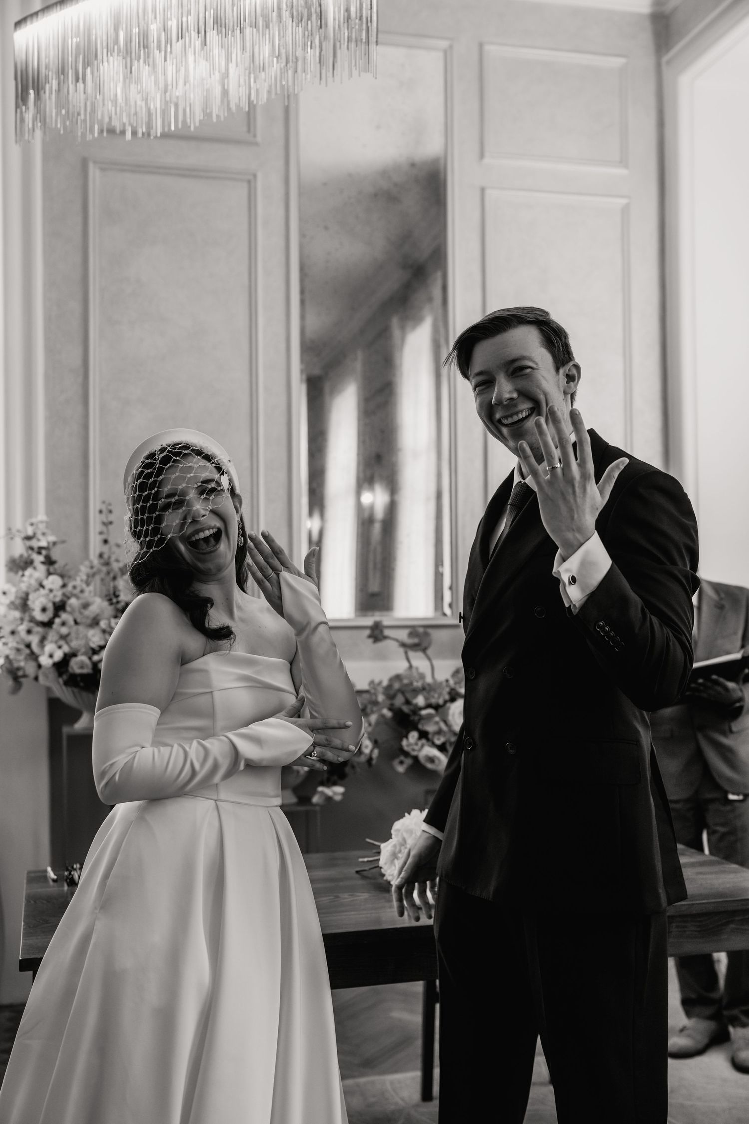 Black and white photo of joyful newlyweds showing wedding rings, bride in strapless gown with veil, groom in dark suit.