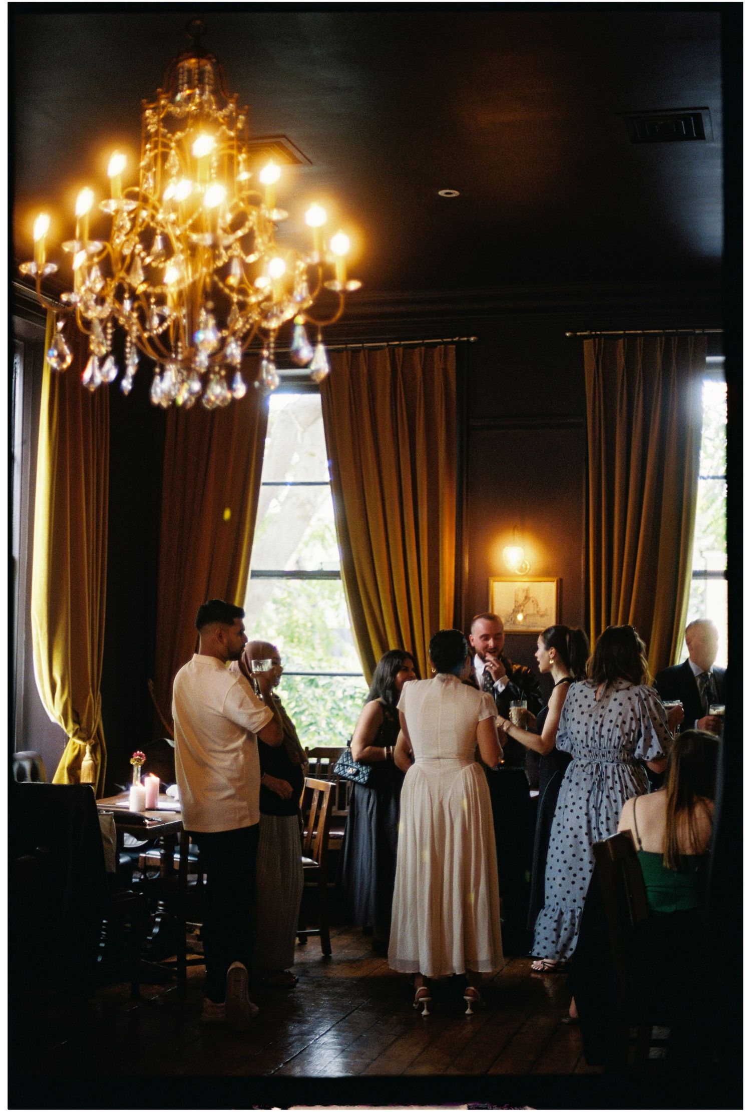 Elegant indoor reception with guests mingling under a crystal chandelier, gold drapes, and warm ambient lighting.