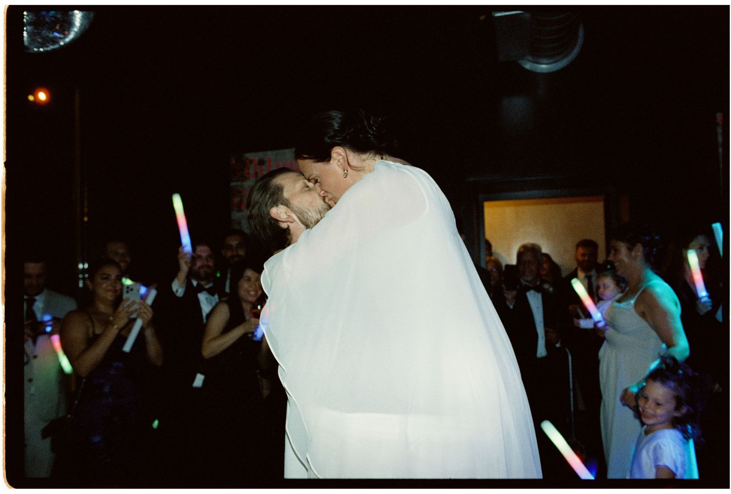 Bride and groom share first dance at wedding reception while guests hold colorful glow sticks in dark ballroom.