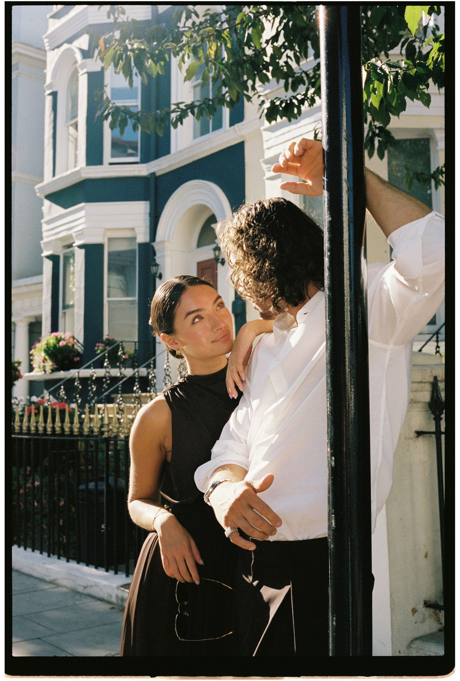 Couple embracing by a black pole on a sunny street with colorful townhouses in the background.